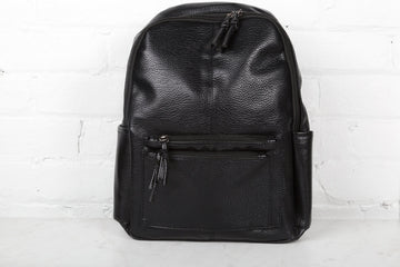 A black backpack with two zipped compartments and a front zip pocket, standing upright against a white brick wall.