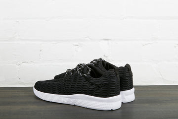 A pair of black low-top lace-up sneakers with white soles, shown from behind against a white brick wall on a dark wood floor.